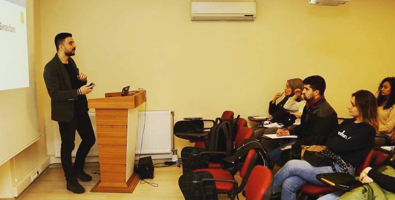 Menteş stands at a podium in front of a projector teaching adults ESL.