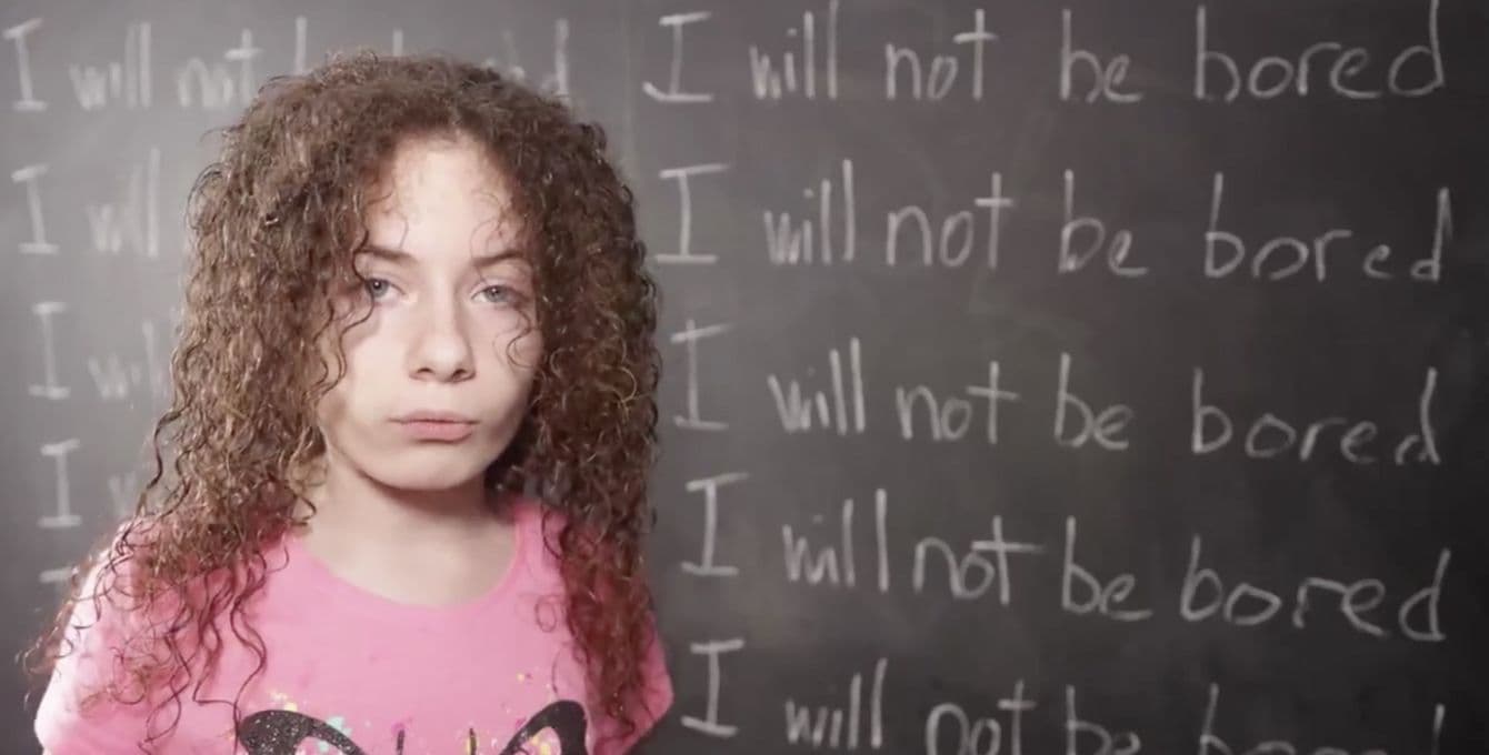 Kid standing in front of chalkboard that says I will not be bored.